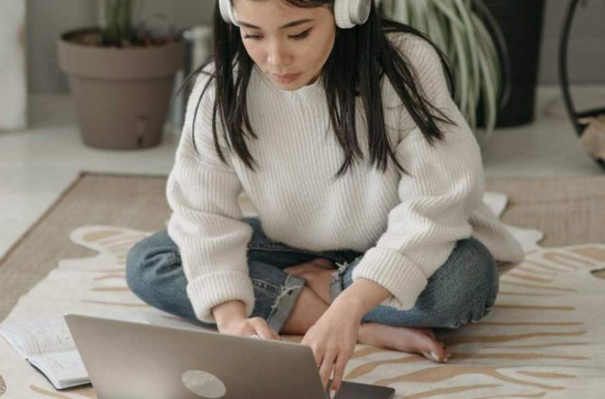 Devojka sa slušalicama sedi na podu i kuca na laptopu.