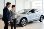 Muškarac i žena u odelima stoje pored srebrnog automobila u salonu